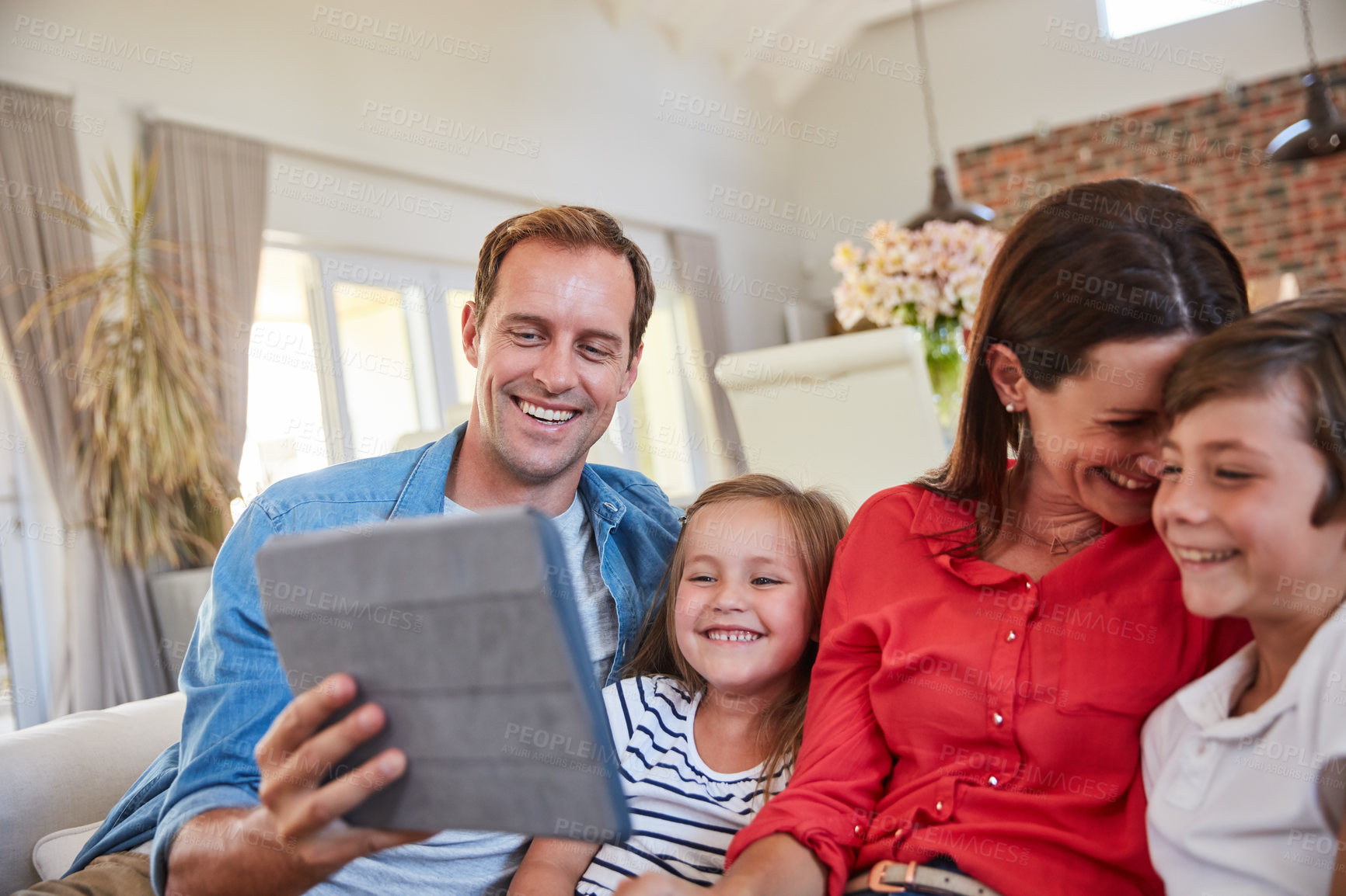 Buy stock photo Shot of smiling parents sitting with their young son and daughter on their living room sofa at home using a digital tablet