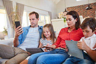 Buy stock photo Shot of a young family sitting together on their living room sofa distracted by various media and devices
