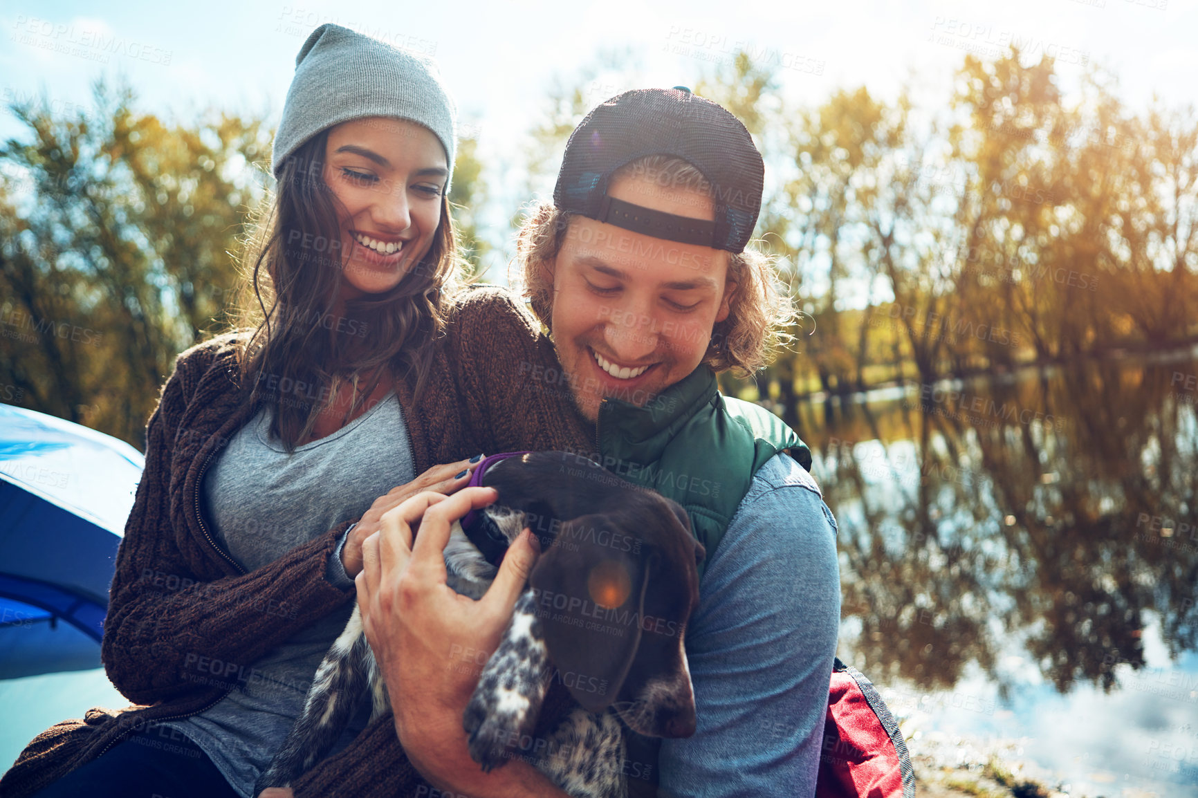 Buy stock photo Smile, camping and couple with dog for adventure by love of freedom in nature lake, sunshine and travel, pet or holiday. Happy, woman and man with puppy for care, hug or relax in summer journey