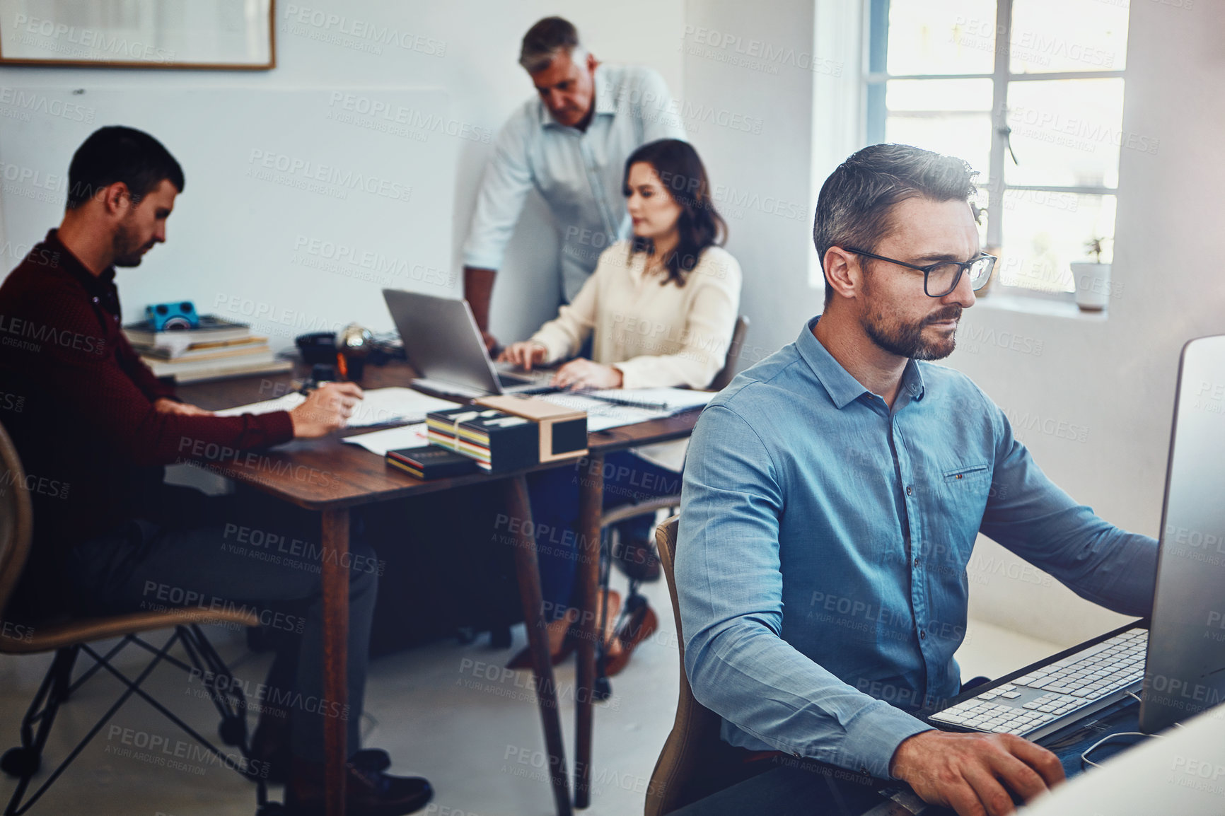 Buy stock photo Shot of a mature man working in the office with his colleagues in the background