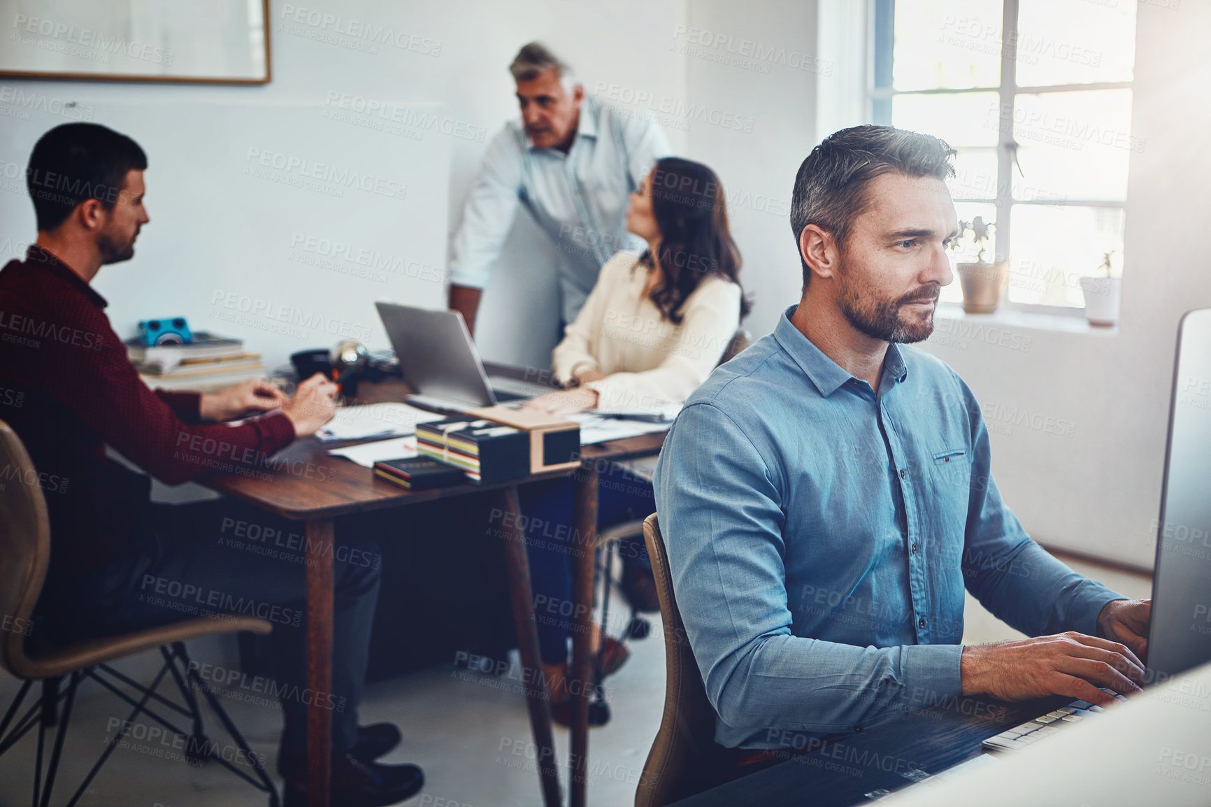 Buy stock photo Shot of a mature man working in the office with his colleagues in the background