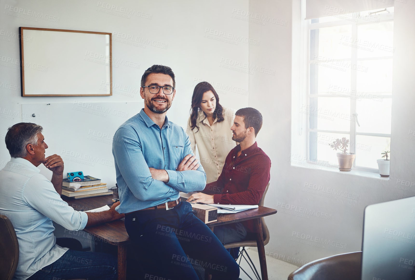 Buy stock photo Portrait of a mature man sitting on a desk in the office which his colleagues are working on