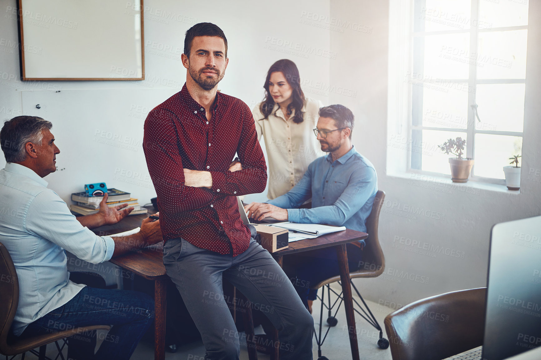 Buy stock photo Portrait of a young man sitting on a desk in the office which his colleagues are working on