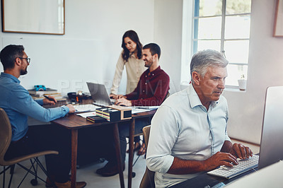 Buy stock photo Shot of a mature man working in the office with his colleagues in the background