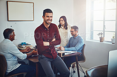 Buy stock photo Portrait of a young man sitting on a desk in the office which his colleagues are working on