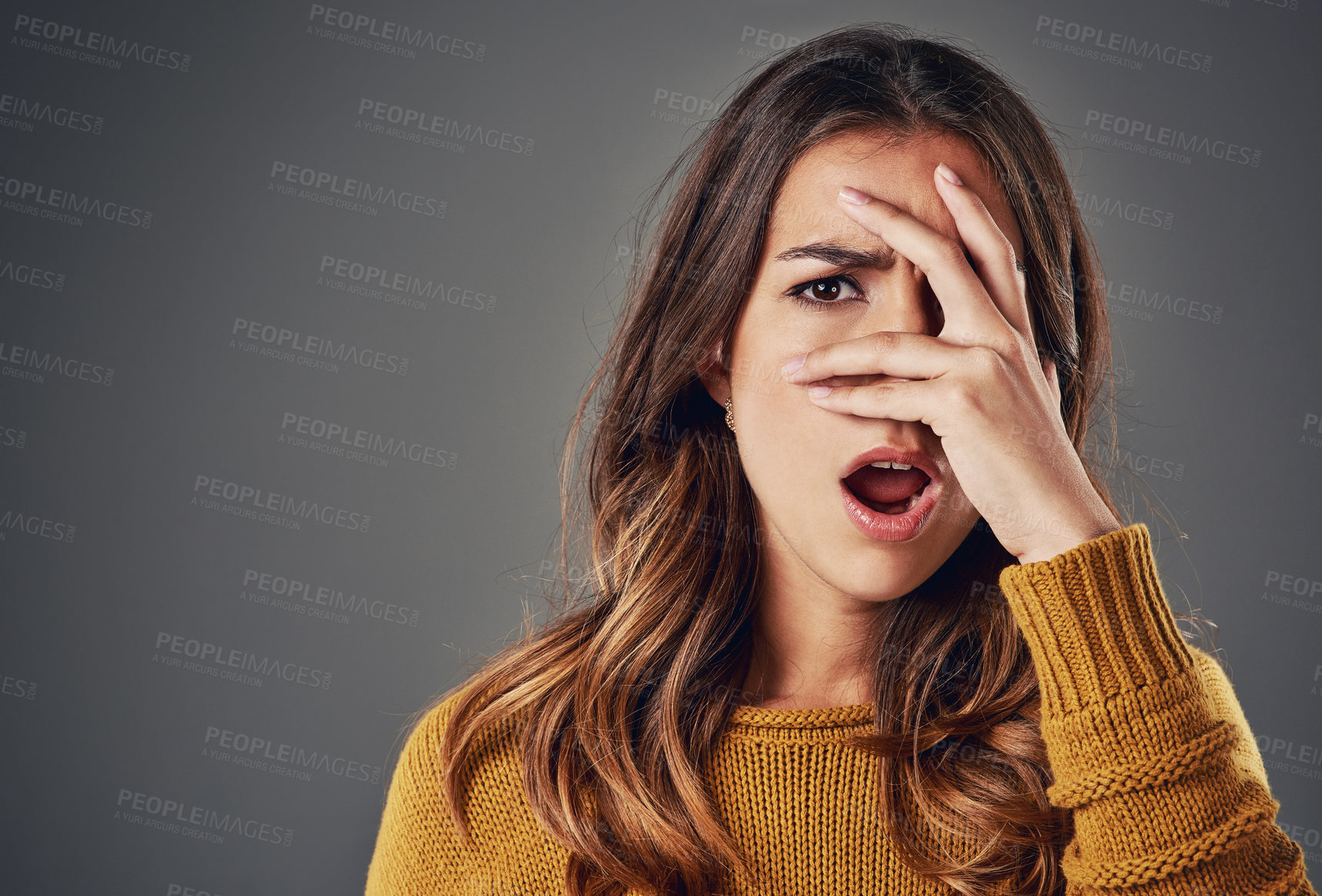 Buy stock photo Portrait of a young woman peeking through her fingers against a grey background