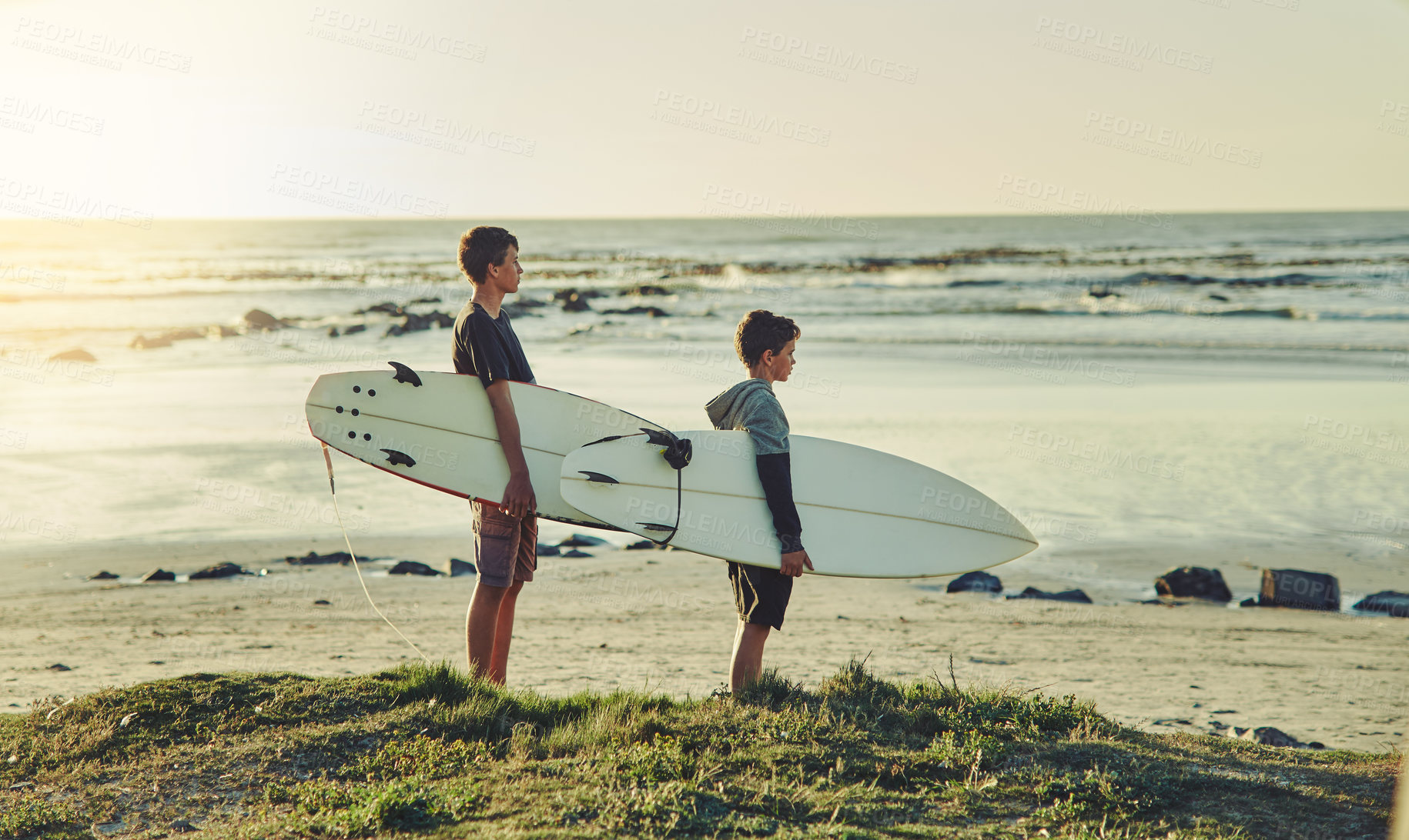 Buy stock photo Shot of two young brothers holding their surfboards while looking towards the ocean