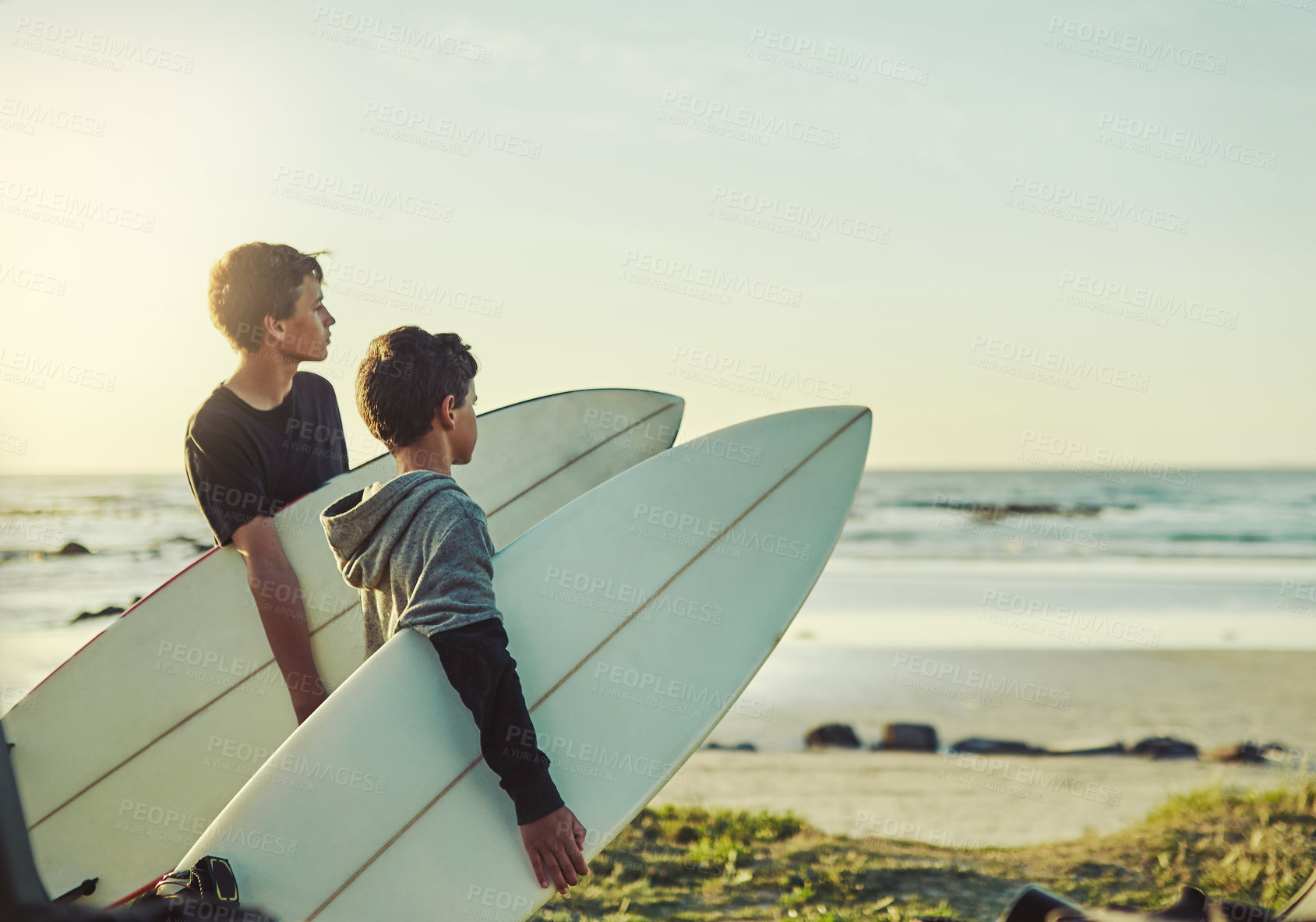 Buy stock photo Shot of two young brothers holding their surfboards while looking towards the ocean