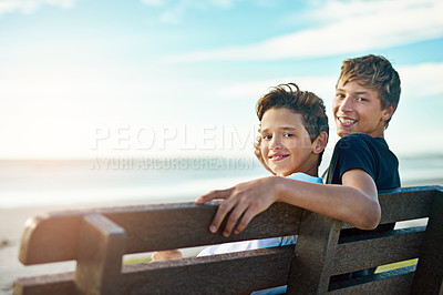 Buy stock photo Portrait of two happy brothers sitting on a bench by the beach together