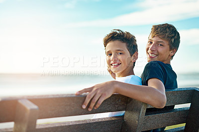 Buy stock photo Portrait of two happy brothers sitting on a bench by the beach together
