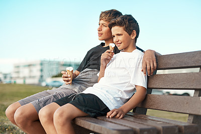 Buy stock photo Shot of two happy brothers eating ice-cream cones while sitting on a bench by the beach