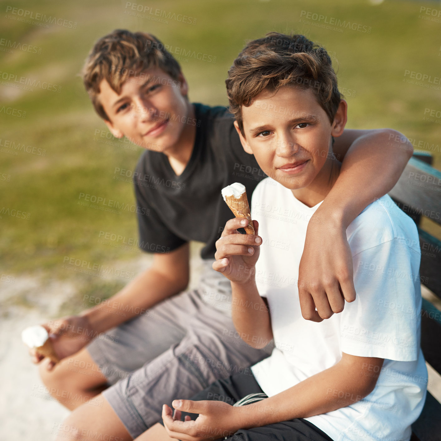 Buy stock photo Portrait of two happy brothers eating ice-cream cones while sitting on a bench by the beach