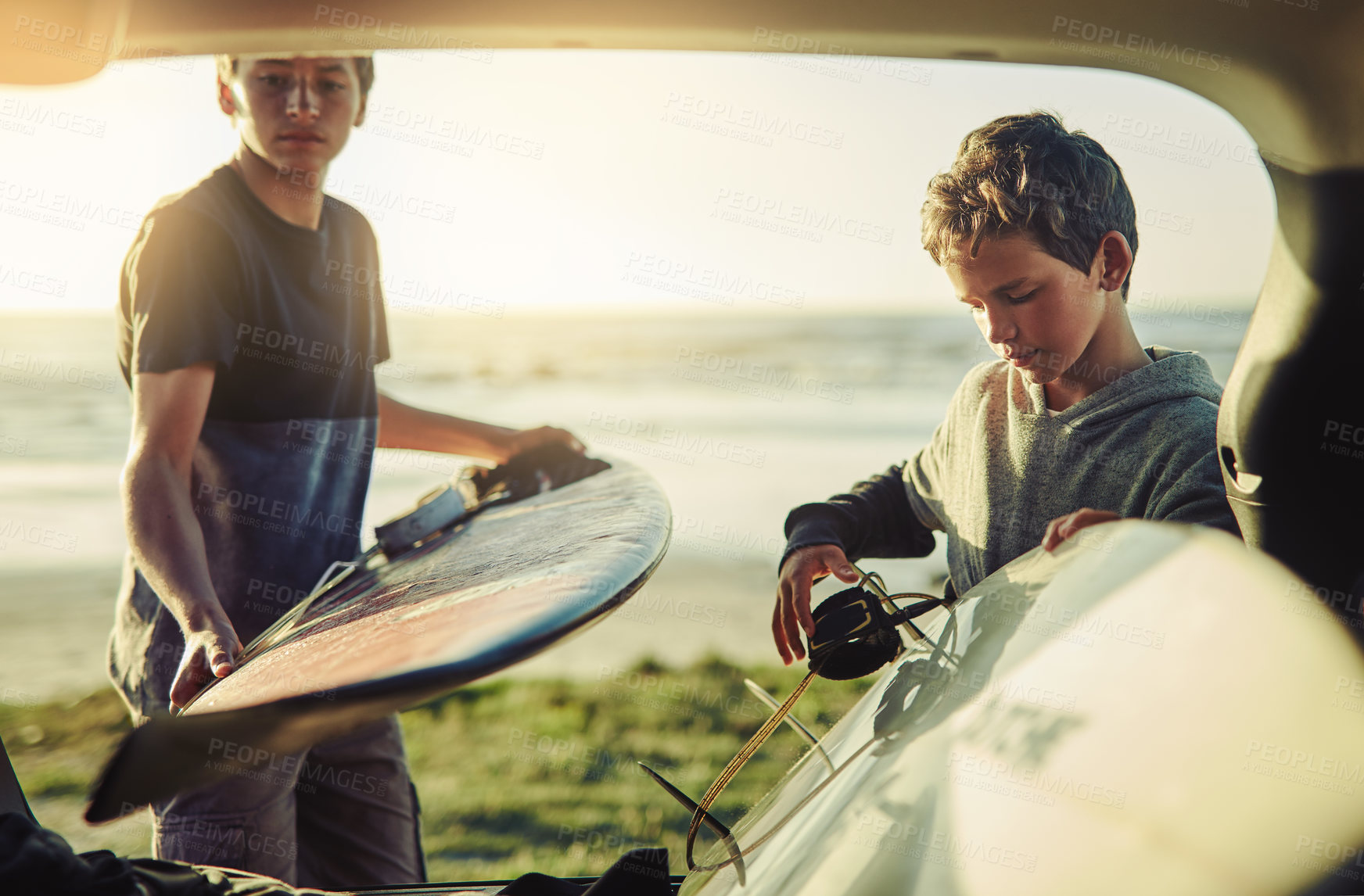 Buy stock photo Shot of two young brothers unloading their surfboards from the back of a car by the beach