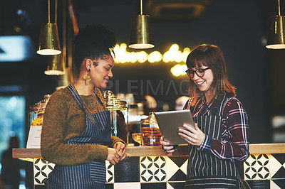 Buy stock photo Shot of two young women using a digital tablet together while working at a coffee shop