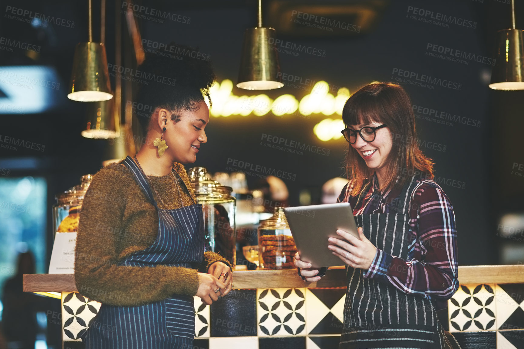 Buy stock photo Shot of two young women using a digital tablet together while working at a coffee shop