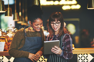 Buy stock photo Shot of two young women using a digital tablet together while working at a coffee shop