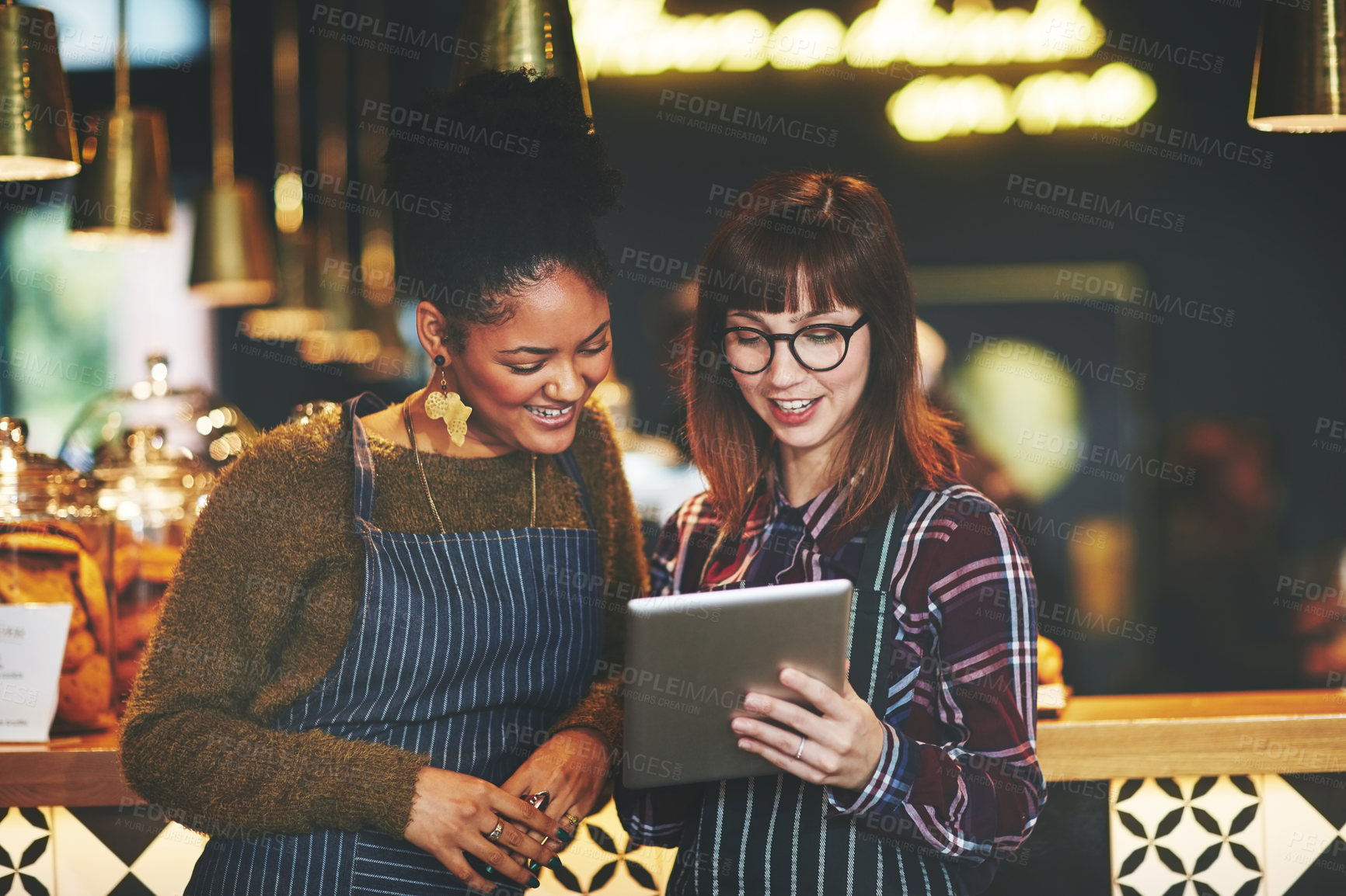 Buy stock photo Shot of two young women using a digital tablet together while working at a coffee shop