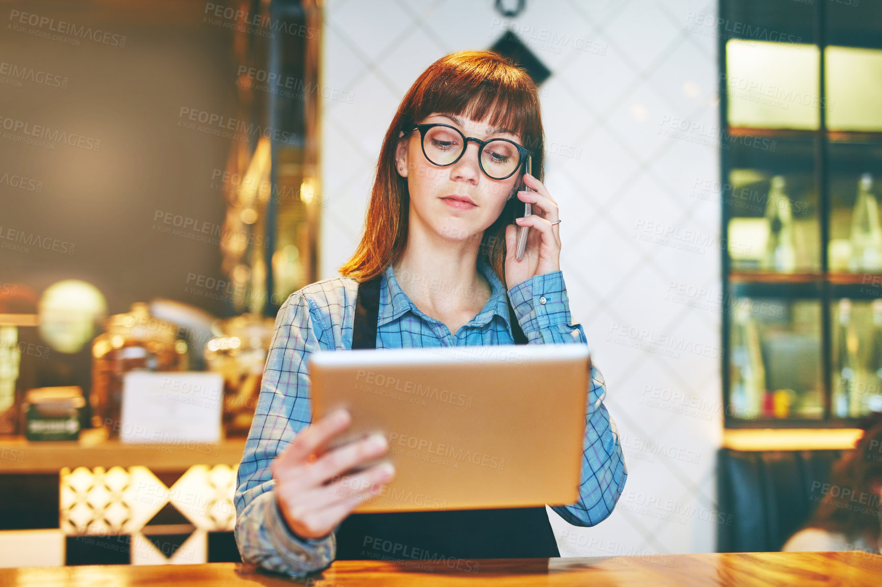 Buy stock photo Shot of a young business owner using a digital tablet while talking on a cellphone in her cafe