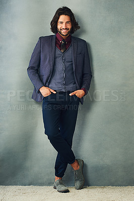 Buy stock photo Studio portrait of a stylishly dressed handsome young man leaning against a gray background with his hand in his pockets