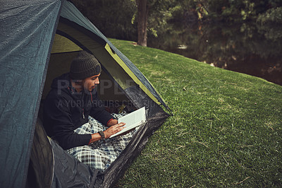 Buy stock photo Shot of a young man reading a book while sitting in his tent at a campsite