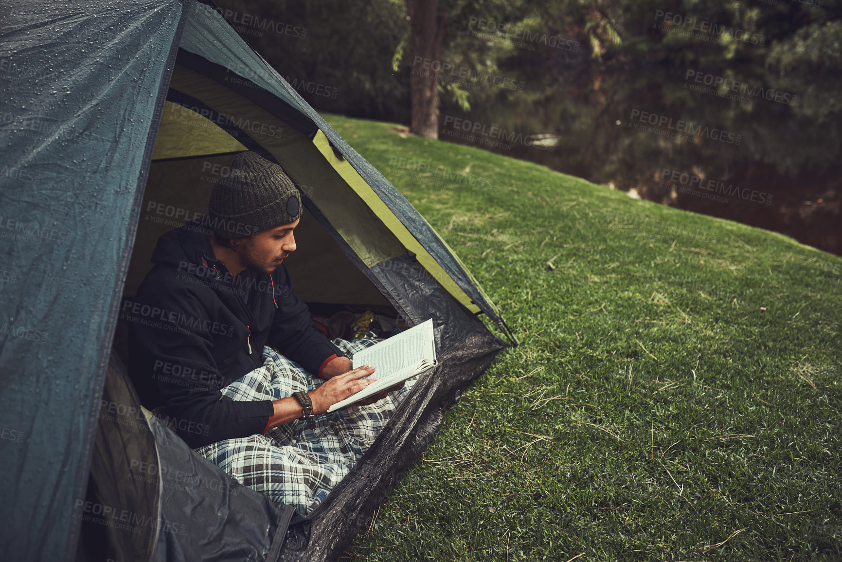 Buy stock photo Shot of a young man reading a book while sitting in his tent at a campsite