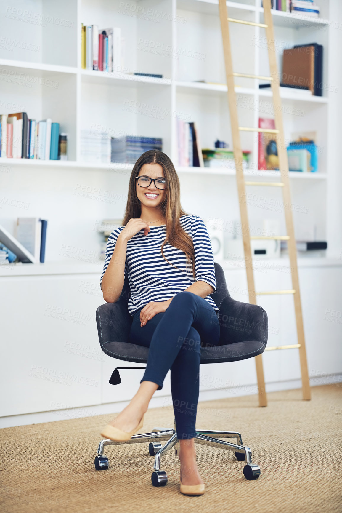 Buy stock photo Portrait of a smiling young woman wearing glasses sitting in front of bookshelves at home