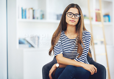 Buy stock photo Portrait of a smiling young woman wearing glasses sitting in front of bookshelves at home