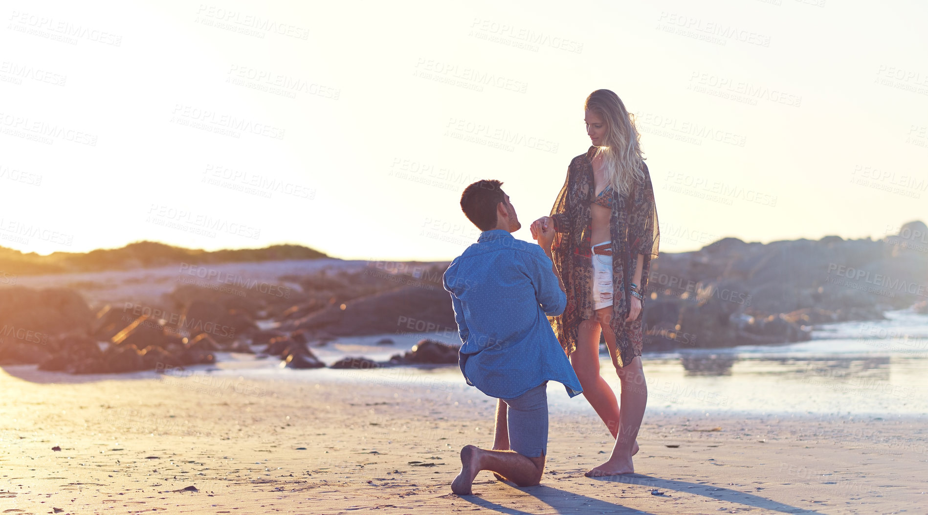 Buy stock photo Shot of a young man holding his girlfriend’s hand on bended knee at the beach