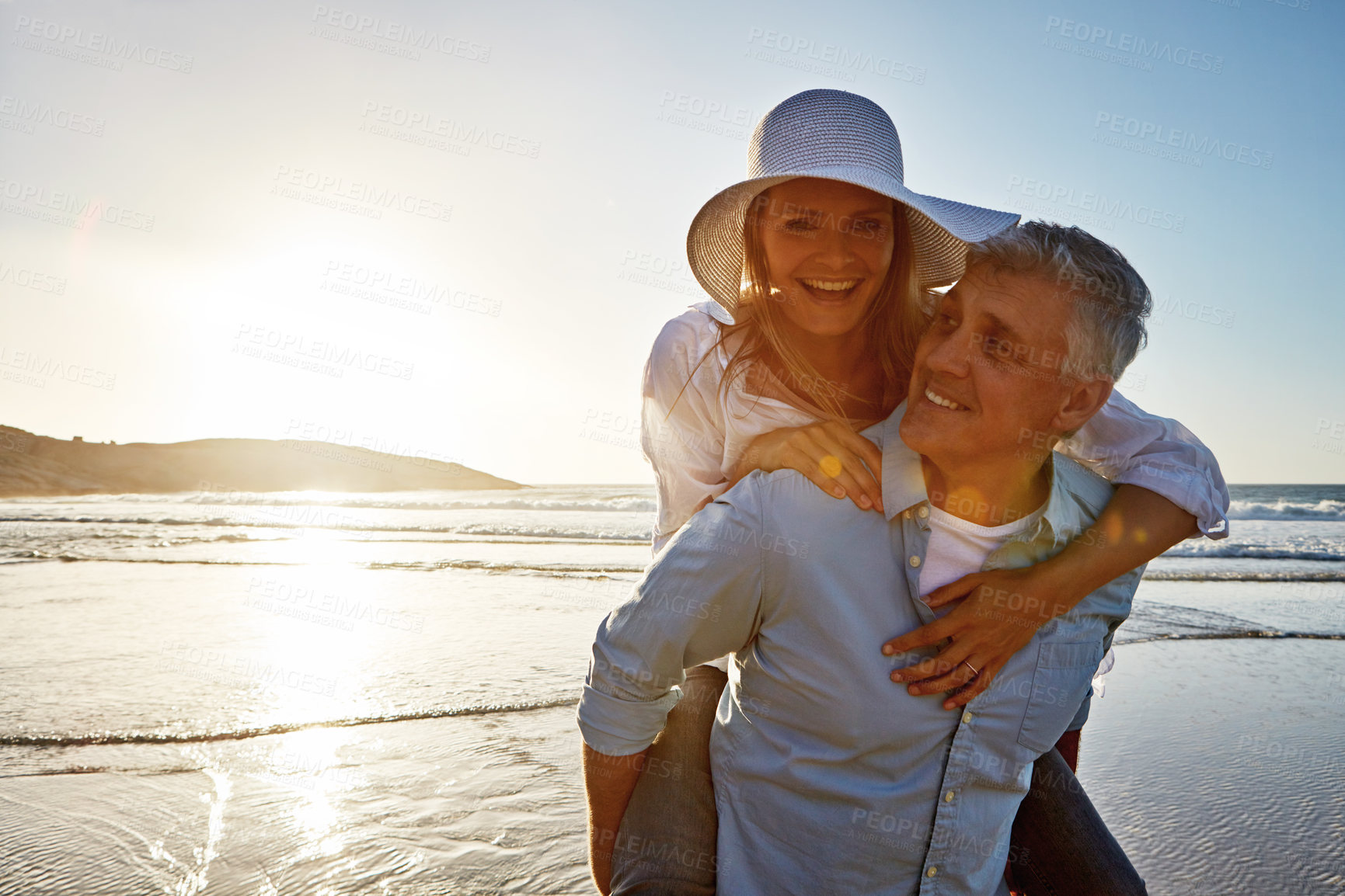 Buy stock photo Mature, happy couple and piggyback with beach sunset for love, embrace or bonding together in nature. Man carrying woman with smile for hug, outdoor summer holiday or sunshine by water or ocean coast