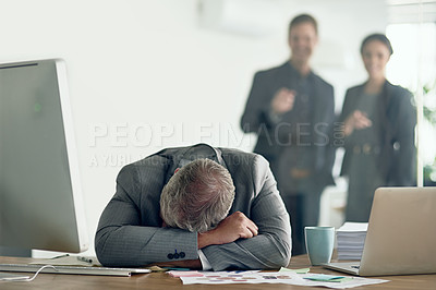 Buy stock photo Shot of a dejected businessman lying on his arms at his desk while his colleagues mock him in the background