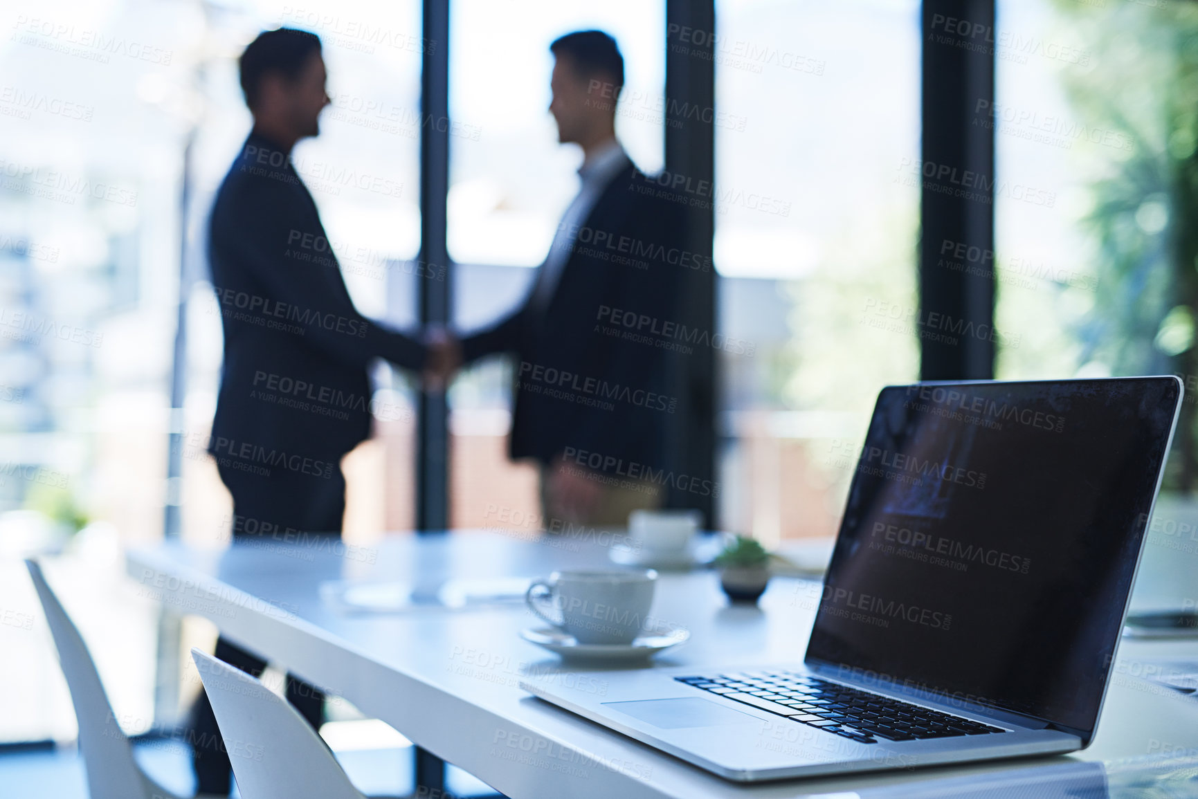 Buy stock photo Shot of a laptop on a table in an office with two businessmen shaking hands in the background
