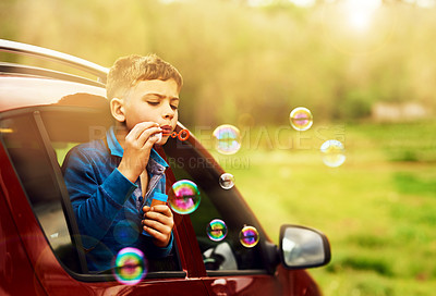 Buy stock photo Shot of a playful little boy blowing bubbles while leaning out of a car window