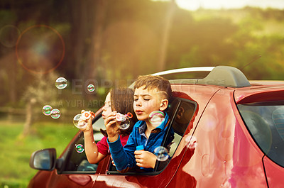 Buy stock photo Shot of two happy children blowing bubbles while leaning out of a car window together