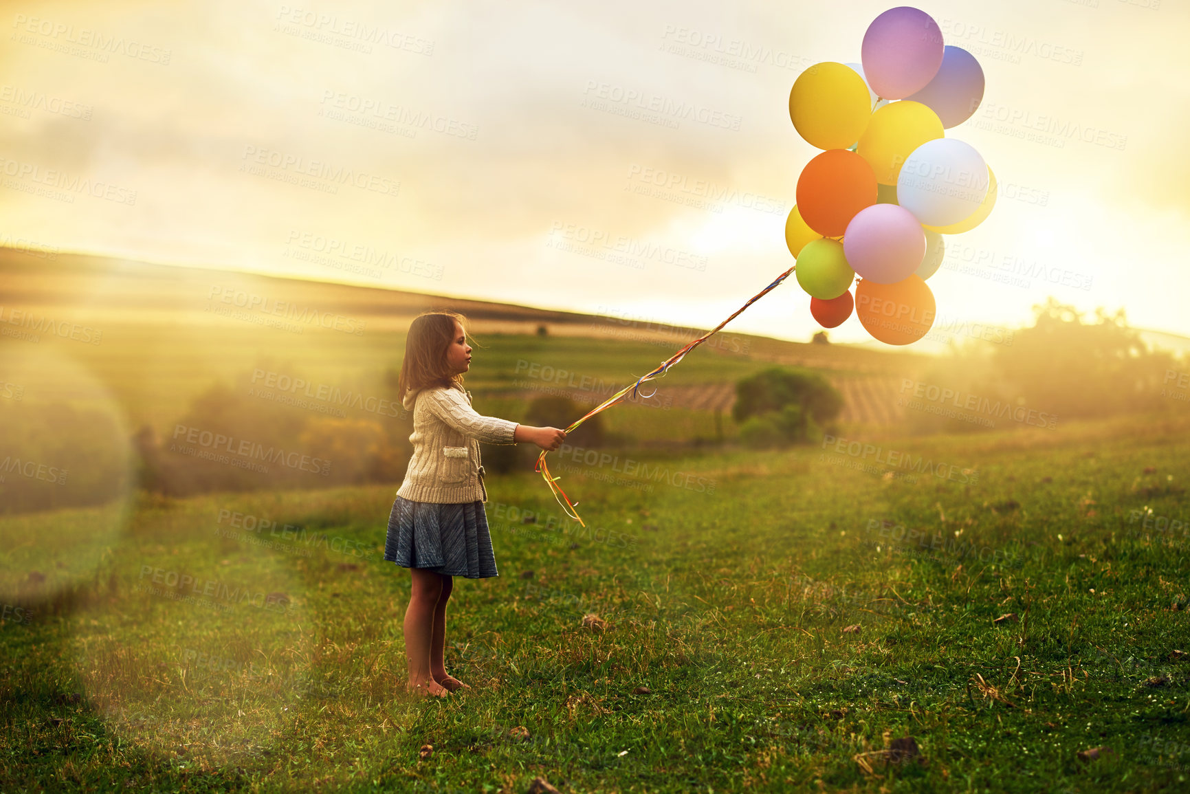 Buy stock photo Shot of a playful little girl walking through a field while holding a bunch of balloons