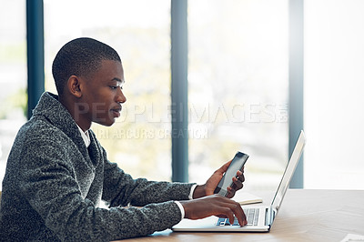 Buy stock photo Shot of a happy young businessman using his smartphone and laptop at a table in the office
