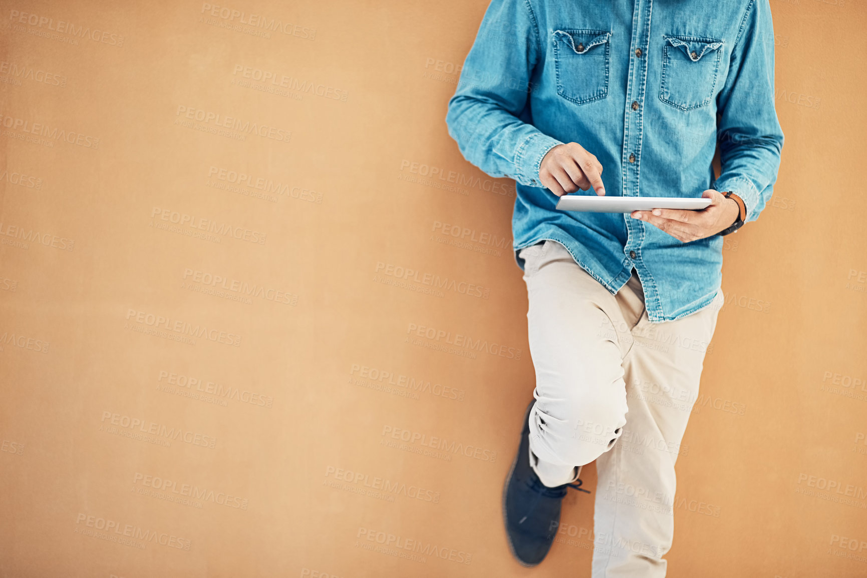 Buy stock photo Shot of an unidentifiable young entrepreneur using his tablet while leaning against a wall in the office