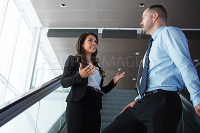 Buy stock photo Shot of two business associates having a conversation while traveling down an escalator in an airport