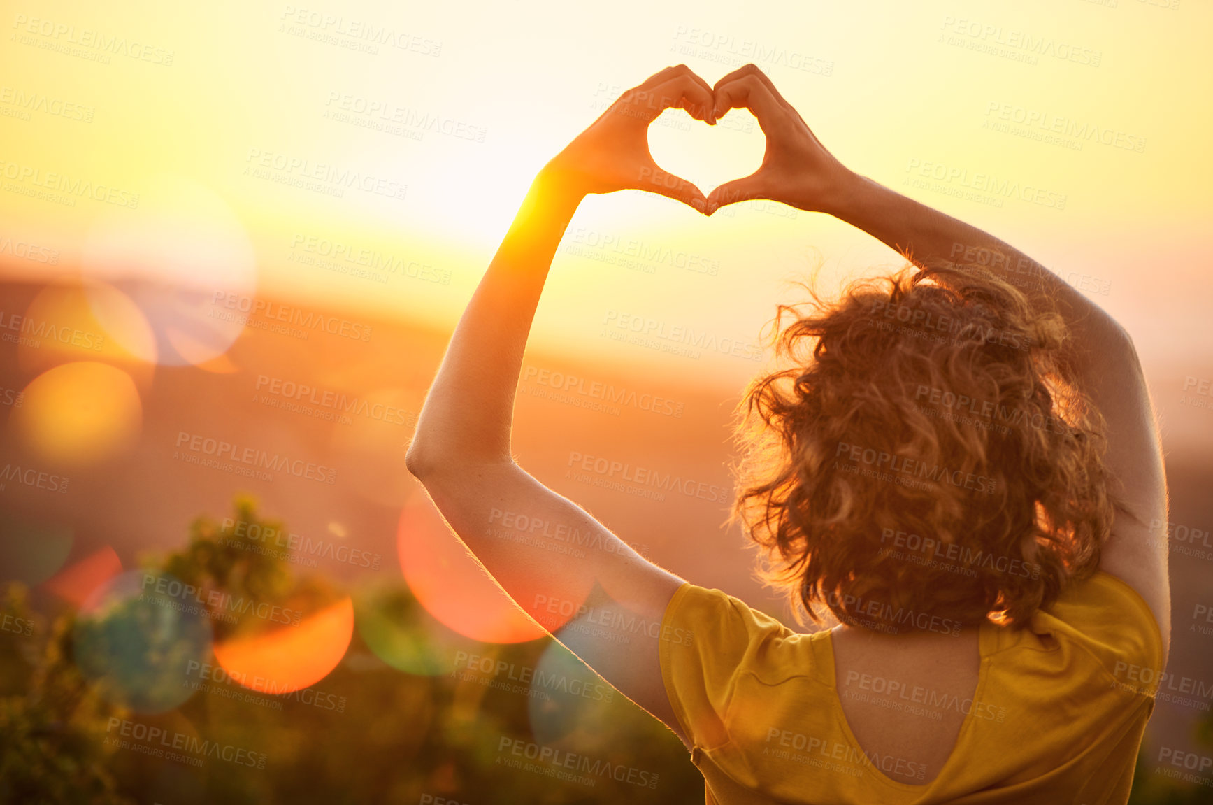 Buy stock photo Rearview shot of an unidentifiable woman making a heart shape with her hands over a sunset landscape
