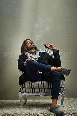 Buy stock photo Shot of a stylishly dressed man smoking an electronic cigarette while sitting in the studio