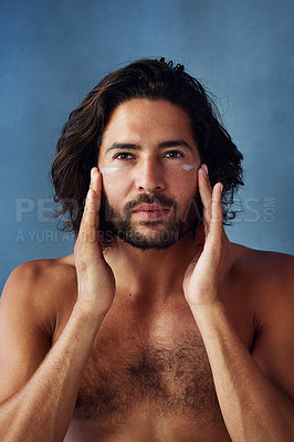 Buy stock photo Studio portrait of a handsome young man applying moisturiser to his face against a blue background