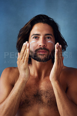 Buy stock photo Studio portrait of a handsome young man applying moisturiser to his face against a blue background