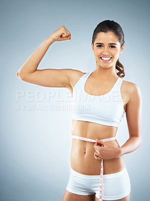 Buy stock photo Studio portrait of an attractive young woman flexing her muscles while measuring her waist against a grey background