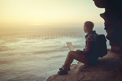 Buy stock photo Full length shot of a young man using his laptop while sitting on a cliff in the mountains