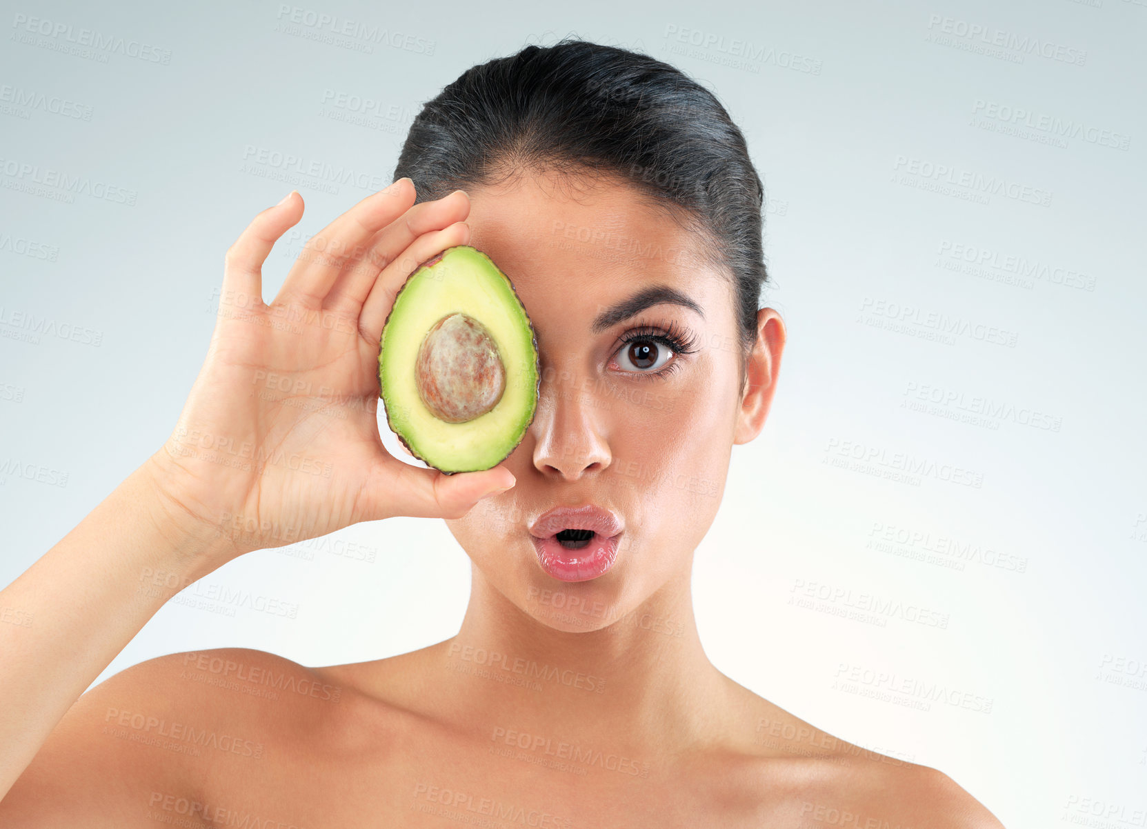 Buy stock photo Studio portrait of a beautiful young woman covering her eye with an avocado against a gray background