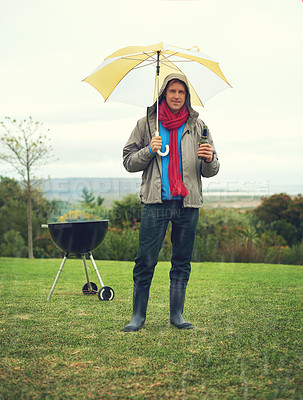 Buy stock photo Shot of a cheerful man barbecuing in the rain while holding an umbrella and drinking a beer