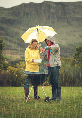 Buy stock photo Shot of a cheerful couple barbecuing in the rain while holding an umbrella and laughing all the way