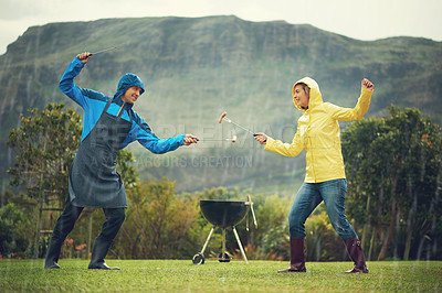 Buy stock photo Shot of a cheerful couple barbecuing in the rain and fencing with barbecue tools and having fun