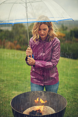 Buy stock photo Shot of a woman having a barbecue outside in the rain while holding an umbrella and looking into the flames
