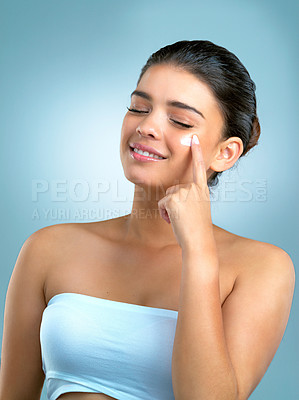 Buy stock photo Studio shot of a beautiful young woman applying moisturiser on her face against a blue background
