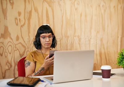 Buy stock photo Shot of a young designer texting on her cellphone while working on a laptop in an office